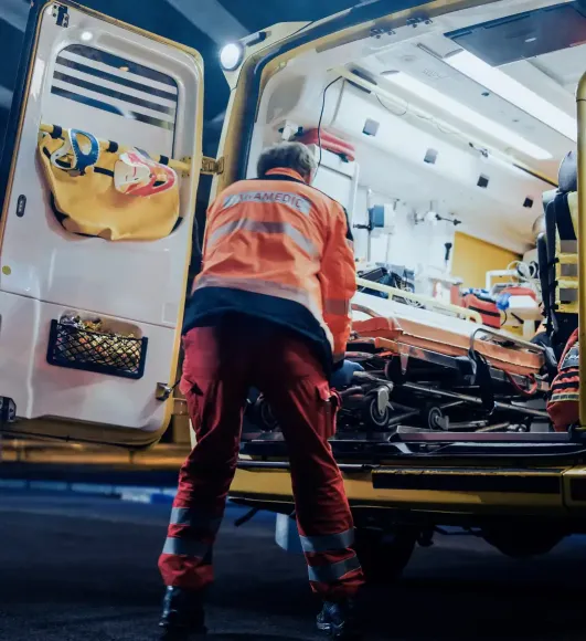 A paramedic loads a stretcher into the back of an ambulance at night, with emergency medical equipment visible inside.
