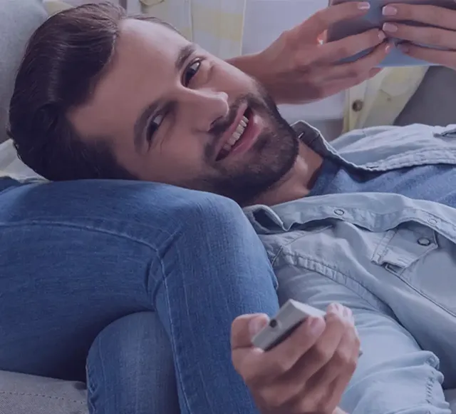 Person reclining on a couch holding a remote, leaning against legs, with a smartphone visible.