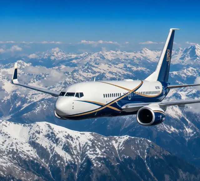 White and blue passenger airplane flying above a snow-covered mountain range under a clear blue sky with clouds in the distance.