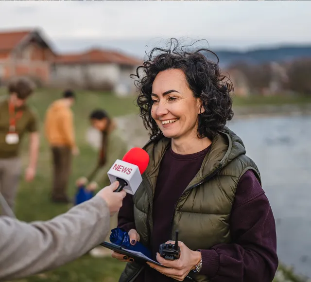Person being interviewed outdoors near a lake, holding recording equipment while a microphone is extended toward them.