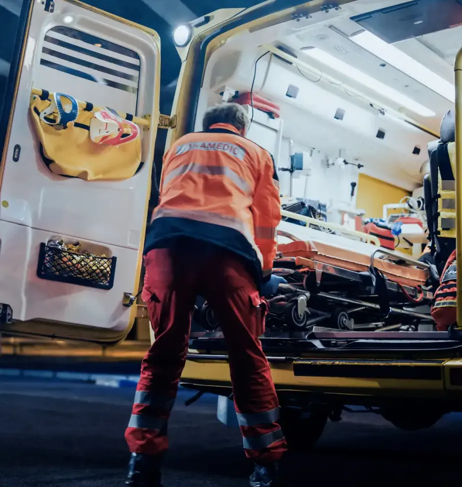 A paramedic loads a stretcher into the back of an ambulance at night, with emergency medical equipment visible inside.