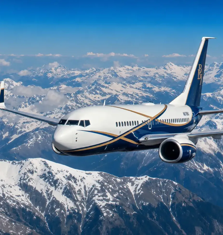 White and blue passenger airplane flying above a snow-covered mountain range under a clear blue sky with clouds in the distance.