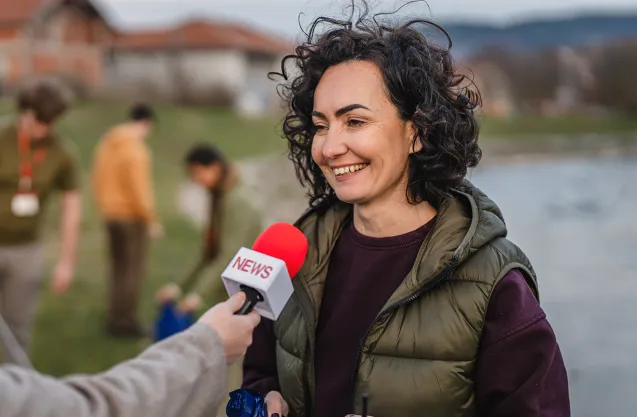 Person being interviewed outdoors near a lake, holding recording equipment while a microphone is extended toward them.