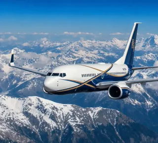 White and blue passenger airplane flying above a snow-covered mountain range under a clear blue sky with clouds in the distance.