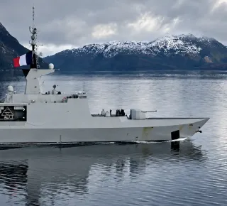 French naval ship on calm water with snow-capped mountains in the background.