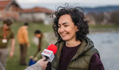 Person being interviewed outdoors near a lake, holding recording equipment while a microphone is extended toward them.